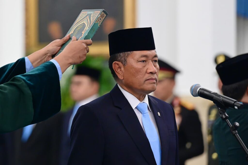 Presidenri go id230720260825326a616d8cf2cb99 39232116 catrawarta A man in a dark suit and light blue tie stands at a formal ceremony while a person in green robes holds up a book over his head a microphone is nearby